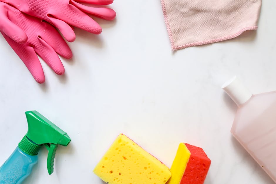 A person wearing bright yellow rubber gloves is cleaning a white tiled wall surface with a blue scrub sponge held in one hand. In the other hand, they are holding a clear spray bottle filled with clea
